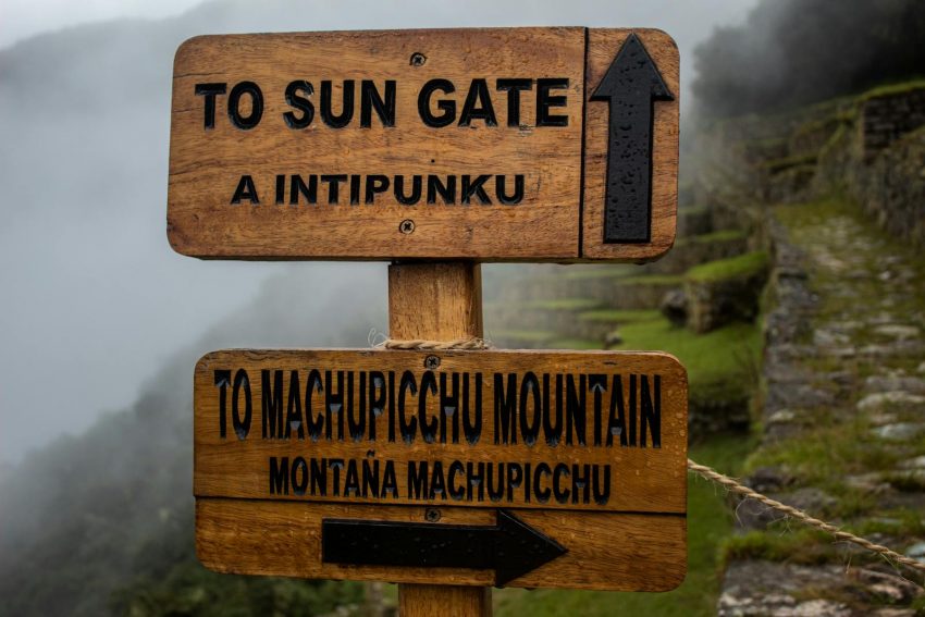 Wooden signpost directing to Sun Gate and Machu Picchu Mountain on misty day.