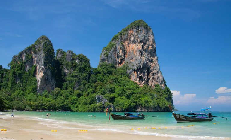 a beach with boats and a large rock formation in the background with Railay Beach in the background, Railay Bay, Thailand