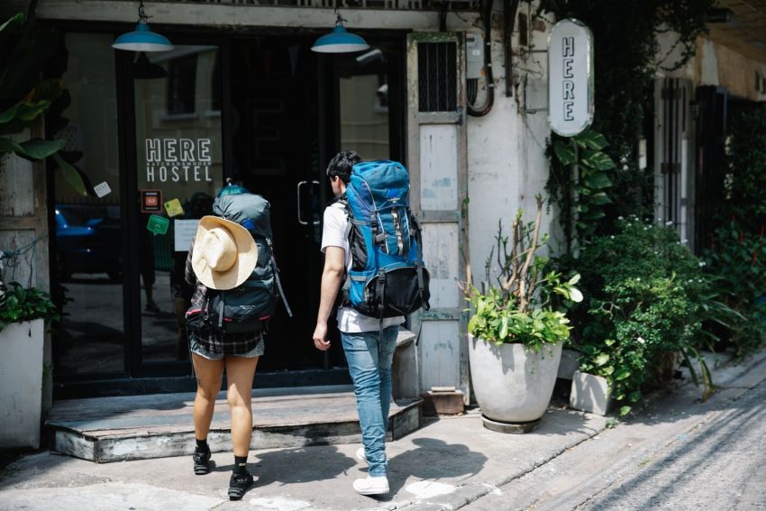 Back view of anonymous couple of tourists with rucksacks walking on street and entering hostel door during trip