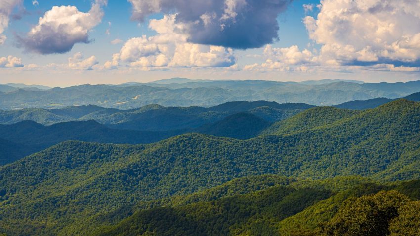 Vast mountain range with lush greenery under a bright blue sky featuring fluffy clouds.